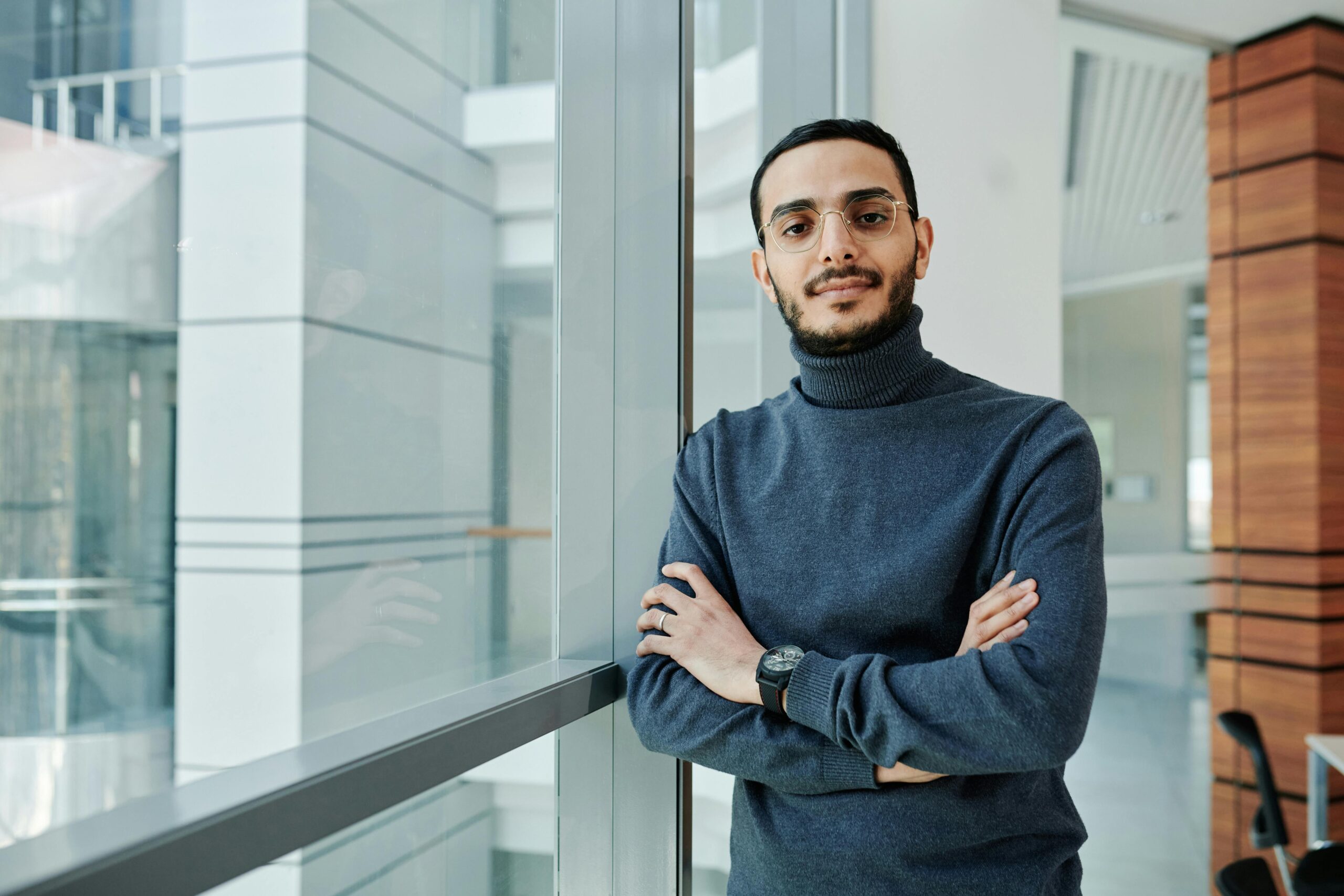 A confident man in a gray turtleneck stands with arms crossed by an office window, exuding professionalism.